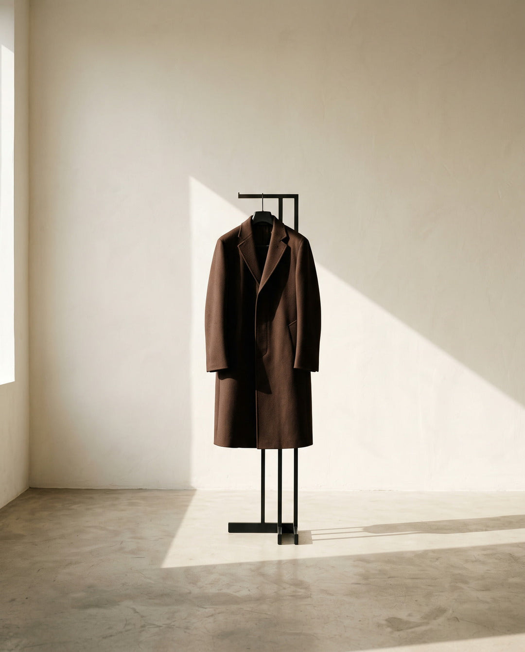 Men's chocolate brown overcoat on a sculptural stand in a bare sunlit room