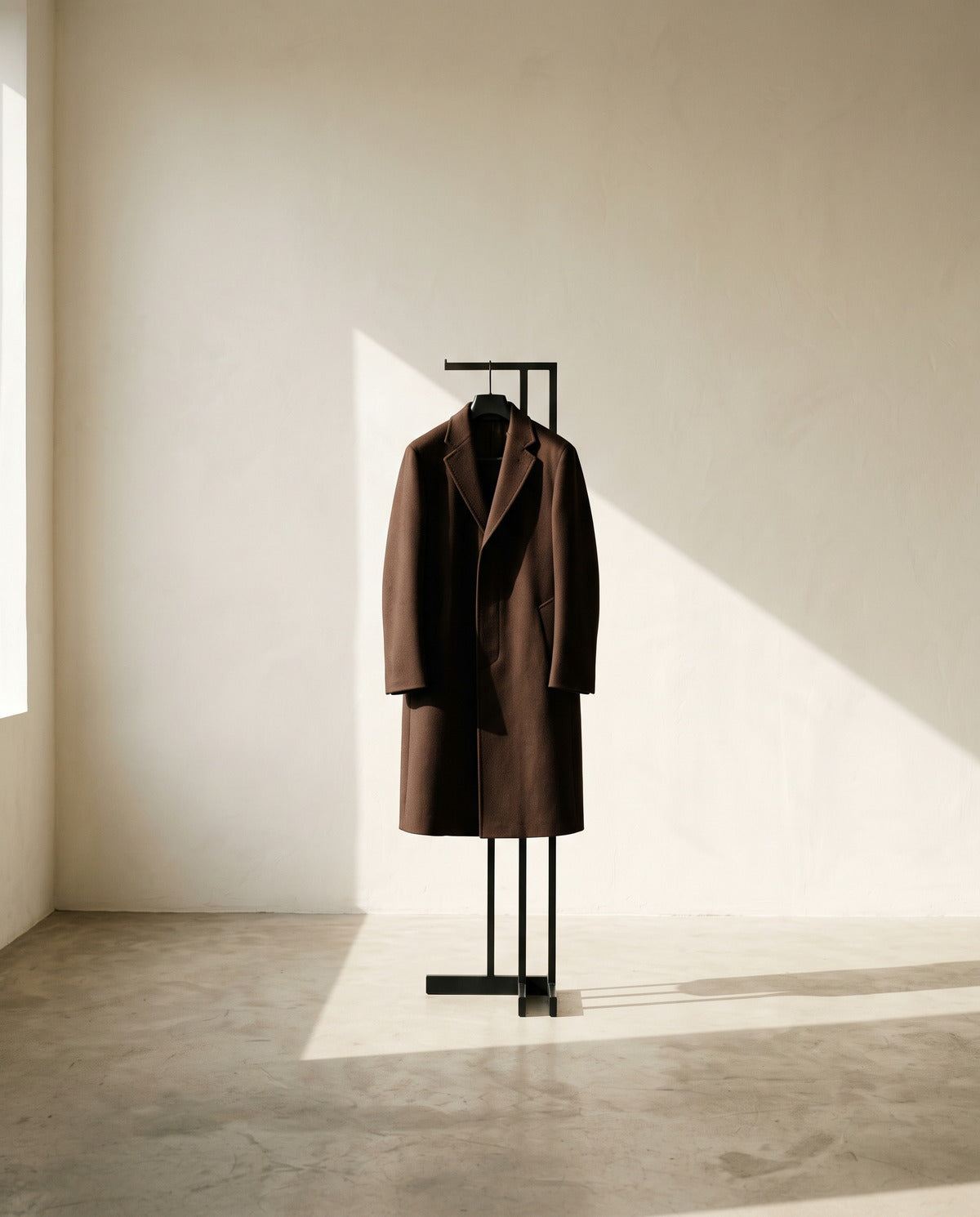 Men's chocolate brown overcoat on a sculptural stand in a bare sunlit room