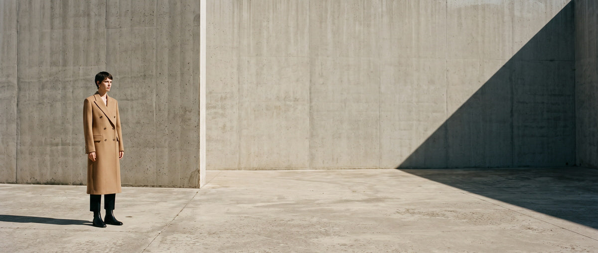 Woman in a long camel wool coat standing in a sunlit concrete courtyard