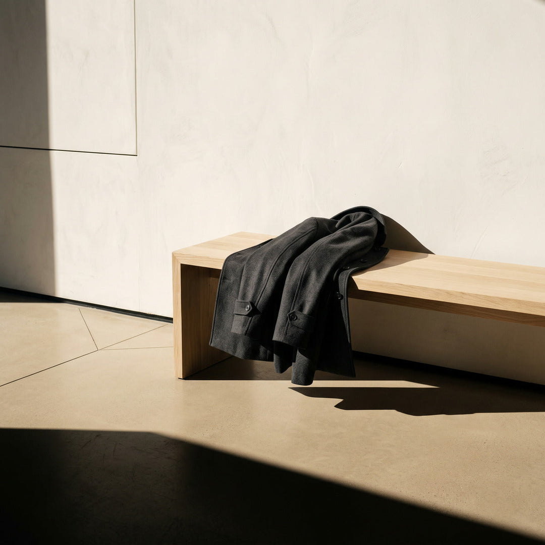 Man's charcoal overcoat draped over a pale wooden bench in raking light